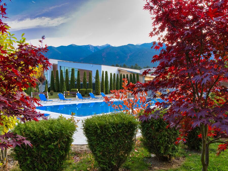 Un edificio moderno con piscina, rodeado de árboles de hojas rojas y montañas al fondo.