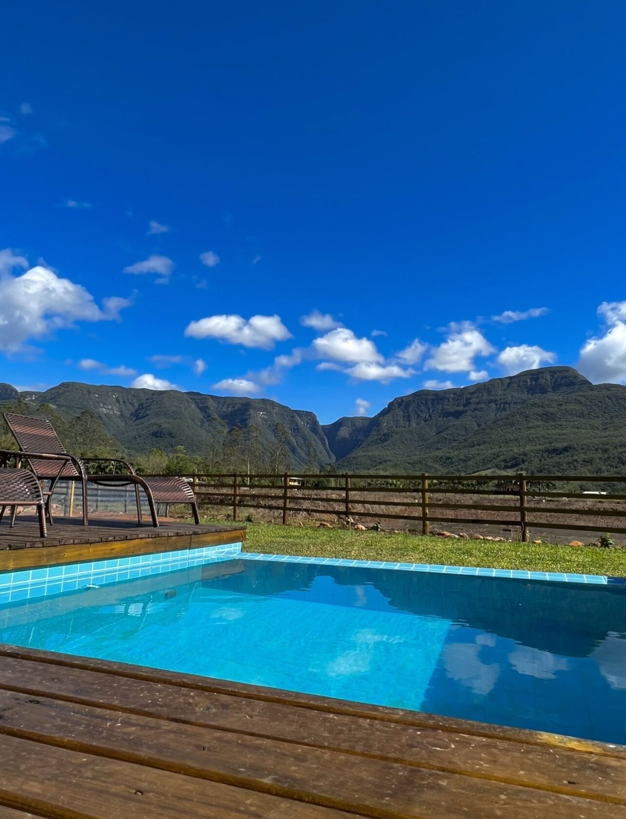 A swimming pool with a wooden deck and lounge chairs, surrounded by a wooden fence, with mountains in the background.