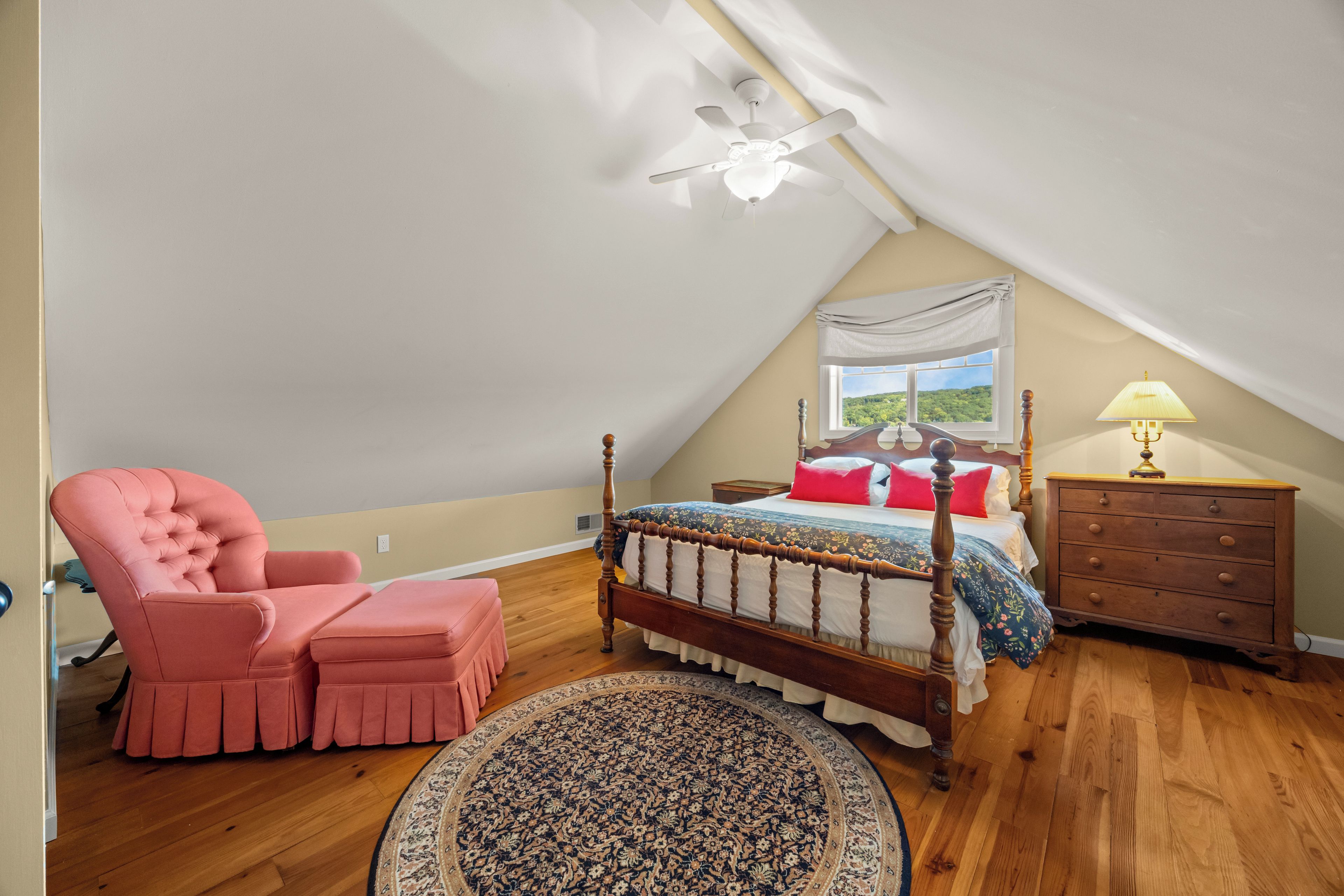 Ein Schlafzimmer mit schrägen Wänden, einem Bett mit Holzgestell, einem rosa Sessel, einer Holzkommode und einem Fenster mit Blick auf Grünflächen.
