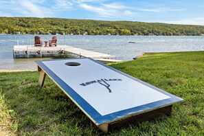 A flat surface with 'KEUKA LAKE' text, a grassy area, a dock with chairs, and a lake with a boat. - Crane Cove: 4 bedroom Lake Home on Keuka Lake (Penn Yan)