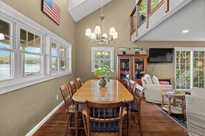A dining area with a wooden table and chairs, a chandelier, and a view of the water through large windows.