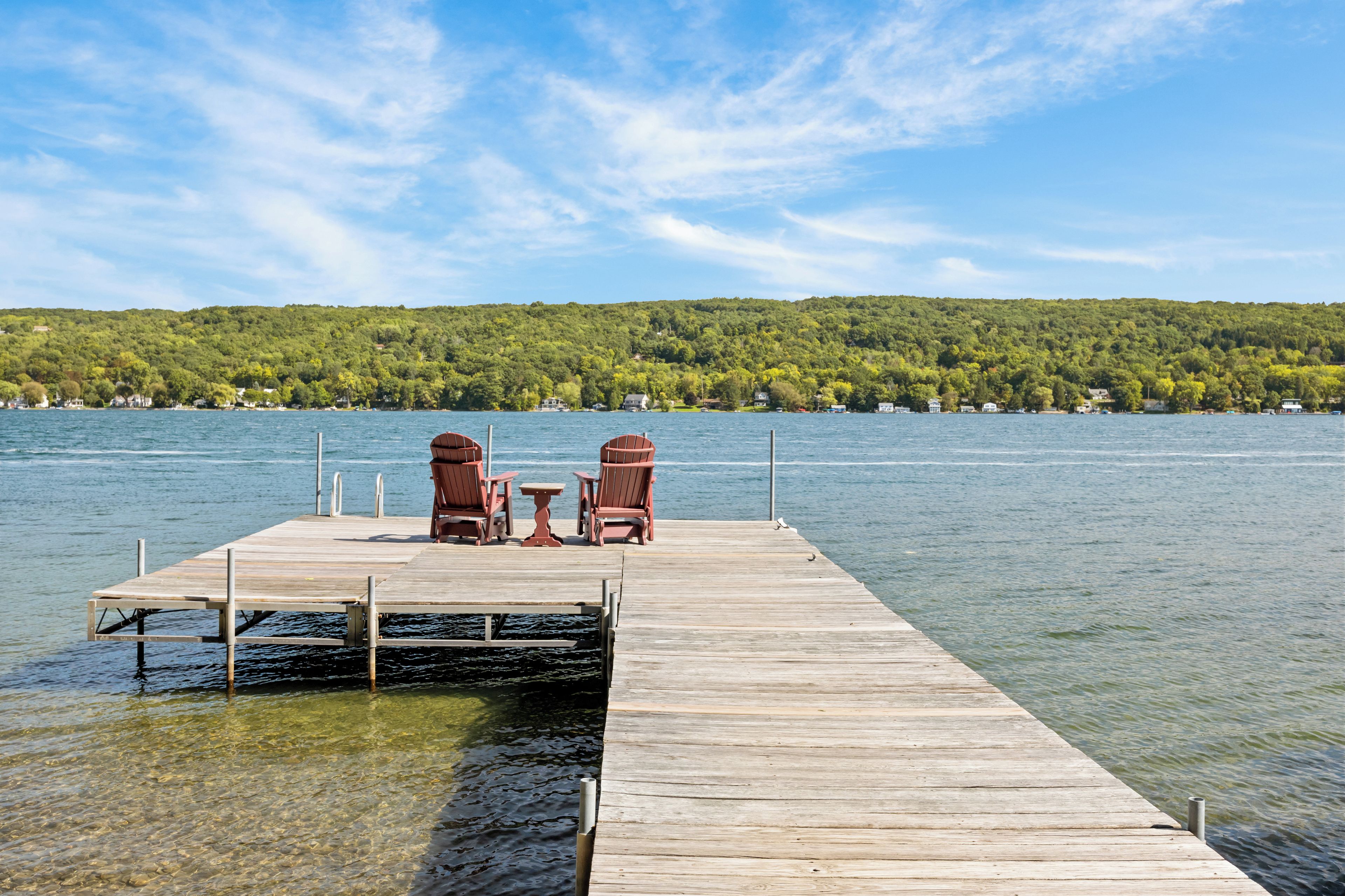 A wooden dock extending into a lake with two Adirondack chairs and a small table, surrounded by a forested hillside.