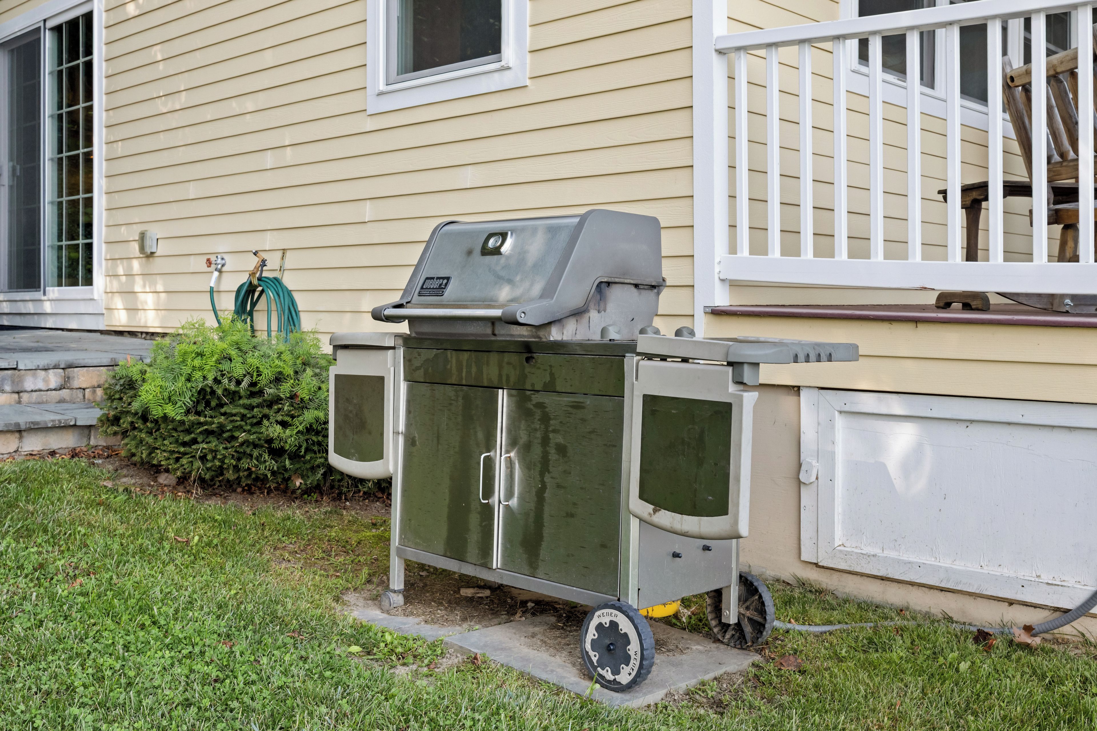 A portable grill unit with wheels, positioned on a concrete pad in front of a house with beige siding and a white porch railing.