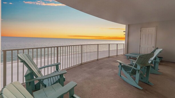 A balcony with a view of the ocean at sunset, featuring two Adirondack chairs and a table. - Gulf Front Luxury-Extra Large Balcony-Regency Isle 907-Signature Properties (Orange Beach)