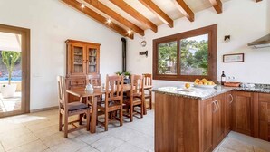 A kitchen with wooden cabinets, a dining table with chairs, a large window, and a countertop with fruits. - Casa Alexa, Strandnähe, Pool, Klimaanlage, Wifi, Ruhige Lage (Cala Sant Vicenç)