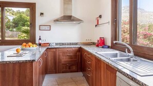 A kitchen with wooden cabinets, granite countertops, a stainless steel range hood, a window with a view of trees, and a sink with a faucet. - Casa Alexa, Strandnähe, Pool, Klimaanlage, Wifi, Ruhige Lage (Cala Sant Vicenç)