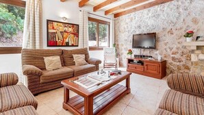 A living room with a striped sofa, a wooden coffee table, a TV, and a stone wall. - Casa Alexa, Strandnähe, Pool, Klimaanlage, Wifi, Ruhige Lage (Cala Sant Vicenç)
