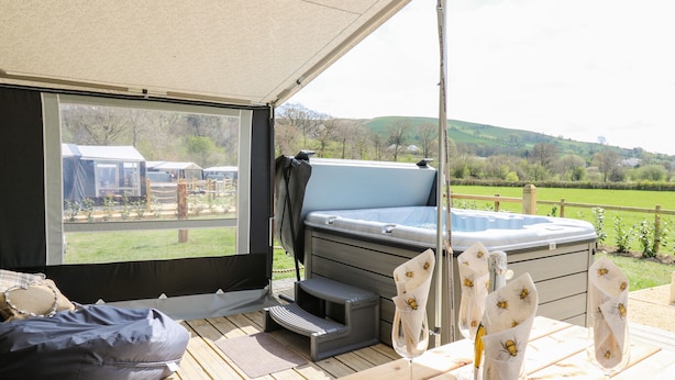 A tented outdoor area with a hot tub, wooden deck, and a view of the countryside.