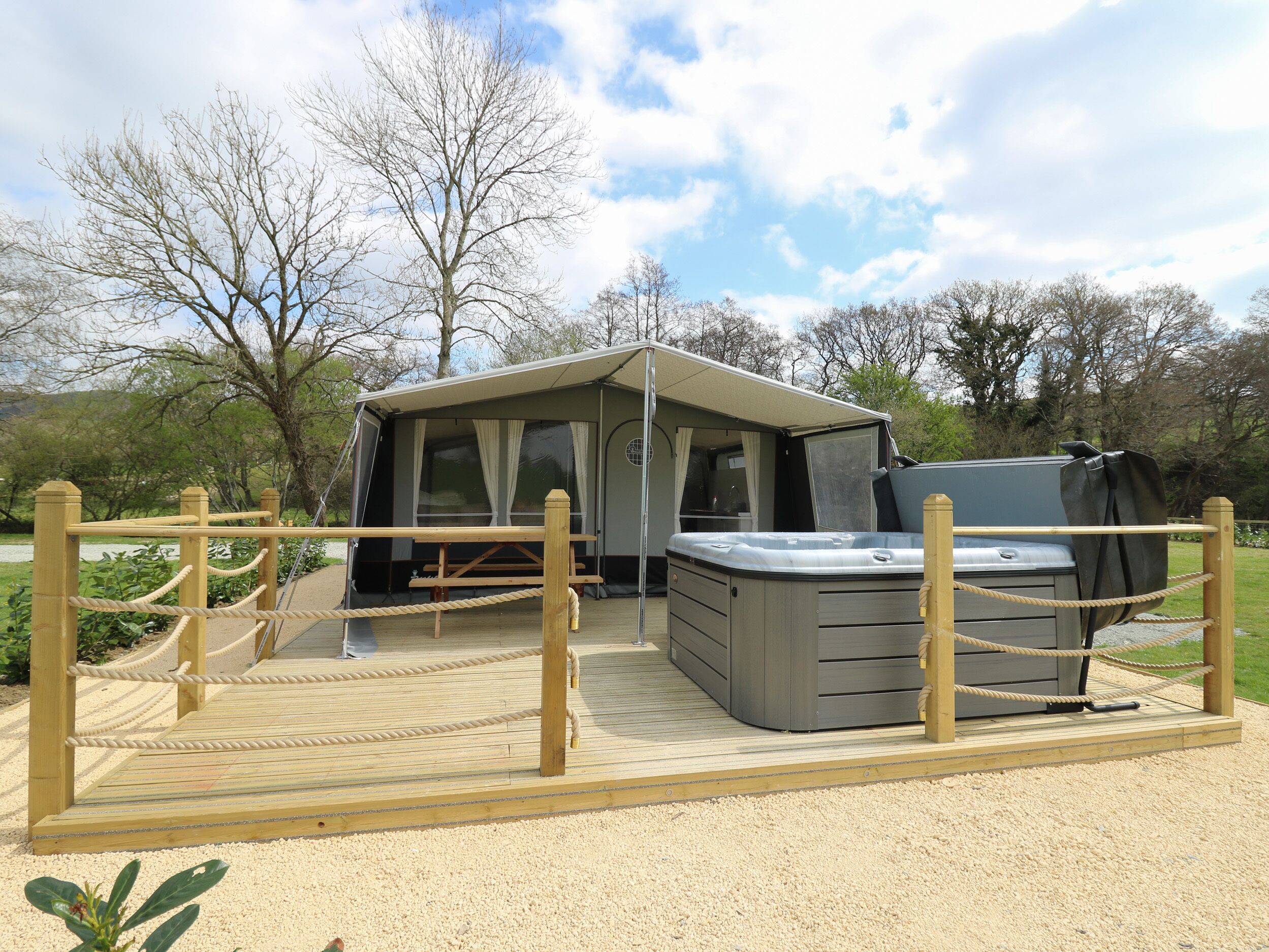 A covered outdoor area with a hot tub and a small building, surrounded by a wooden fence and gravel.