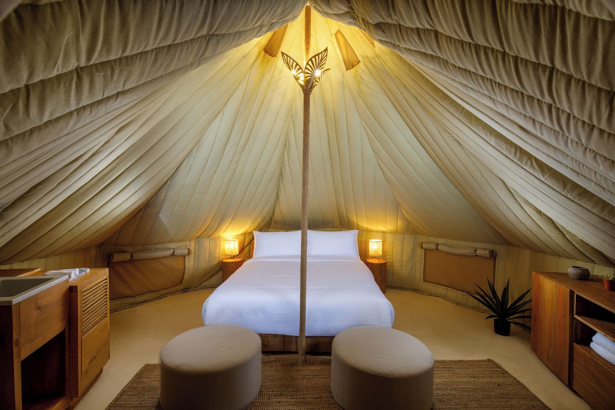 A cozy tented accommodation with a central bed, bedside lamps, and a small seating area.