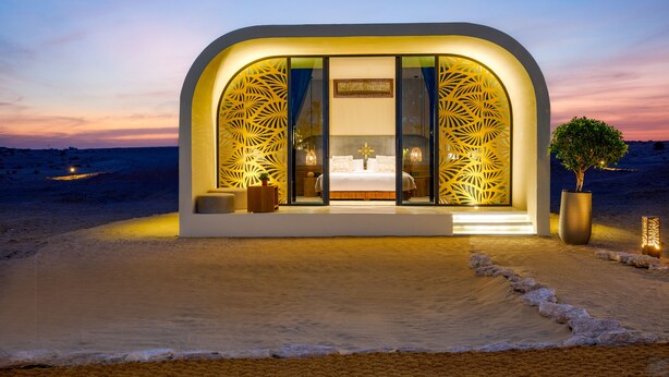 A modern, arched structure with a large window and decorative panels, situated on a beach with a view of the sea and a sunset sky.