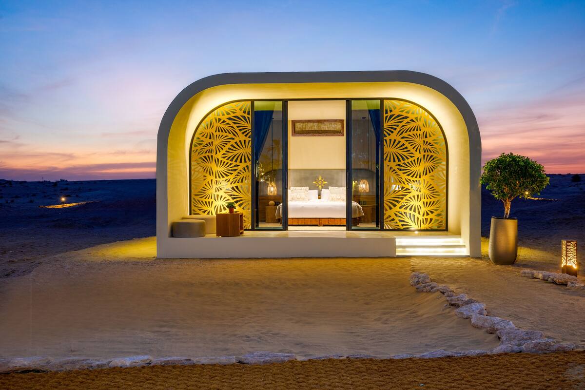 A modern, arched structure with a large window and decorative panels, situated on a beach with a view of the sea and a sunset sky.