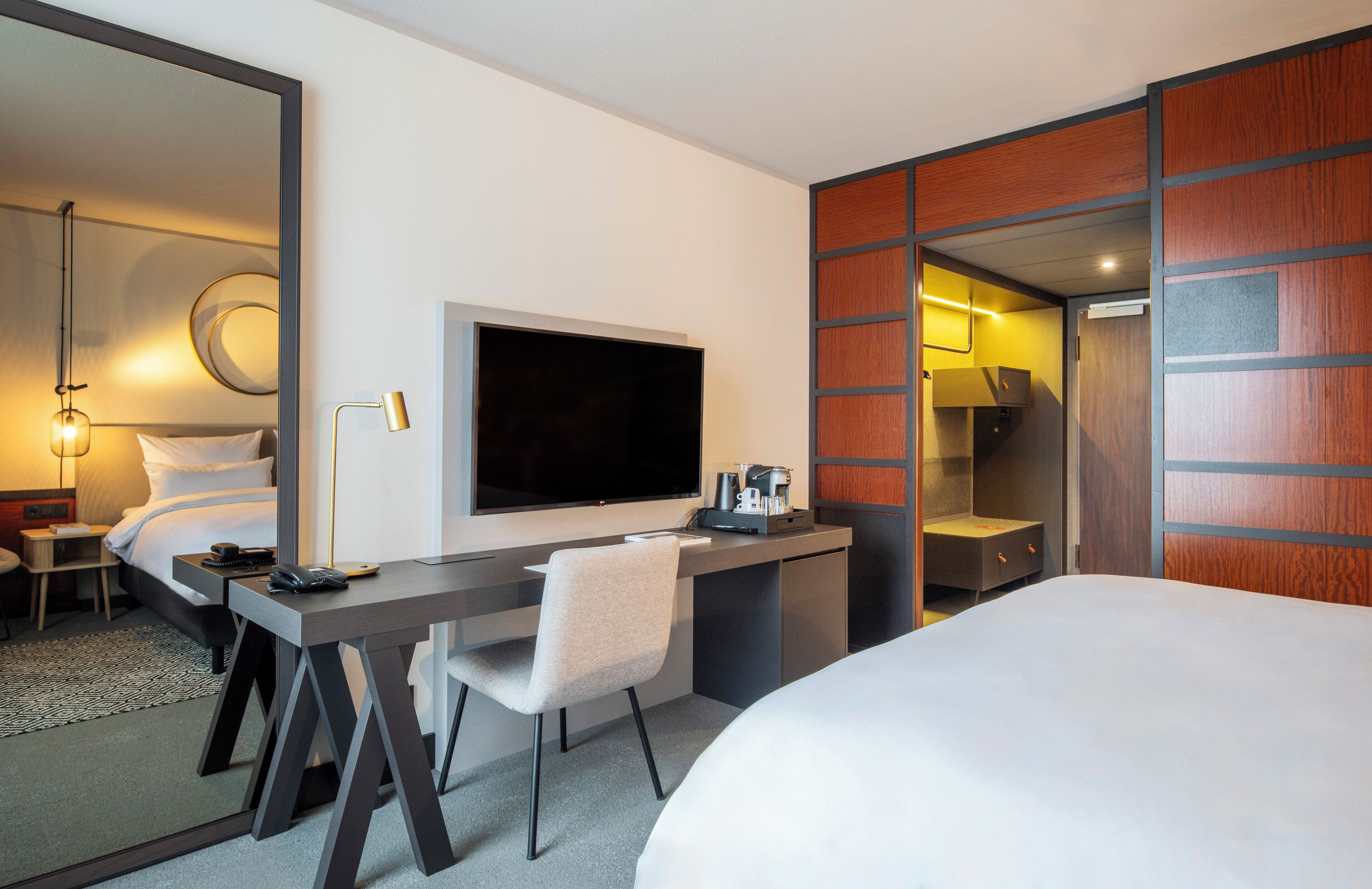 Een moderne hotelkamer met een groot bed, een bureau met stoel, een flatscreen-tv, een koffiezetapparaat en een garderobe met spiegel.