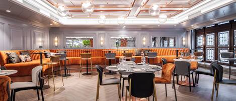 A modern dining area with a mix of seating options, including a long orange sofa and individual chairs, arranged around a central round table.