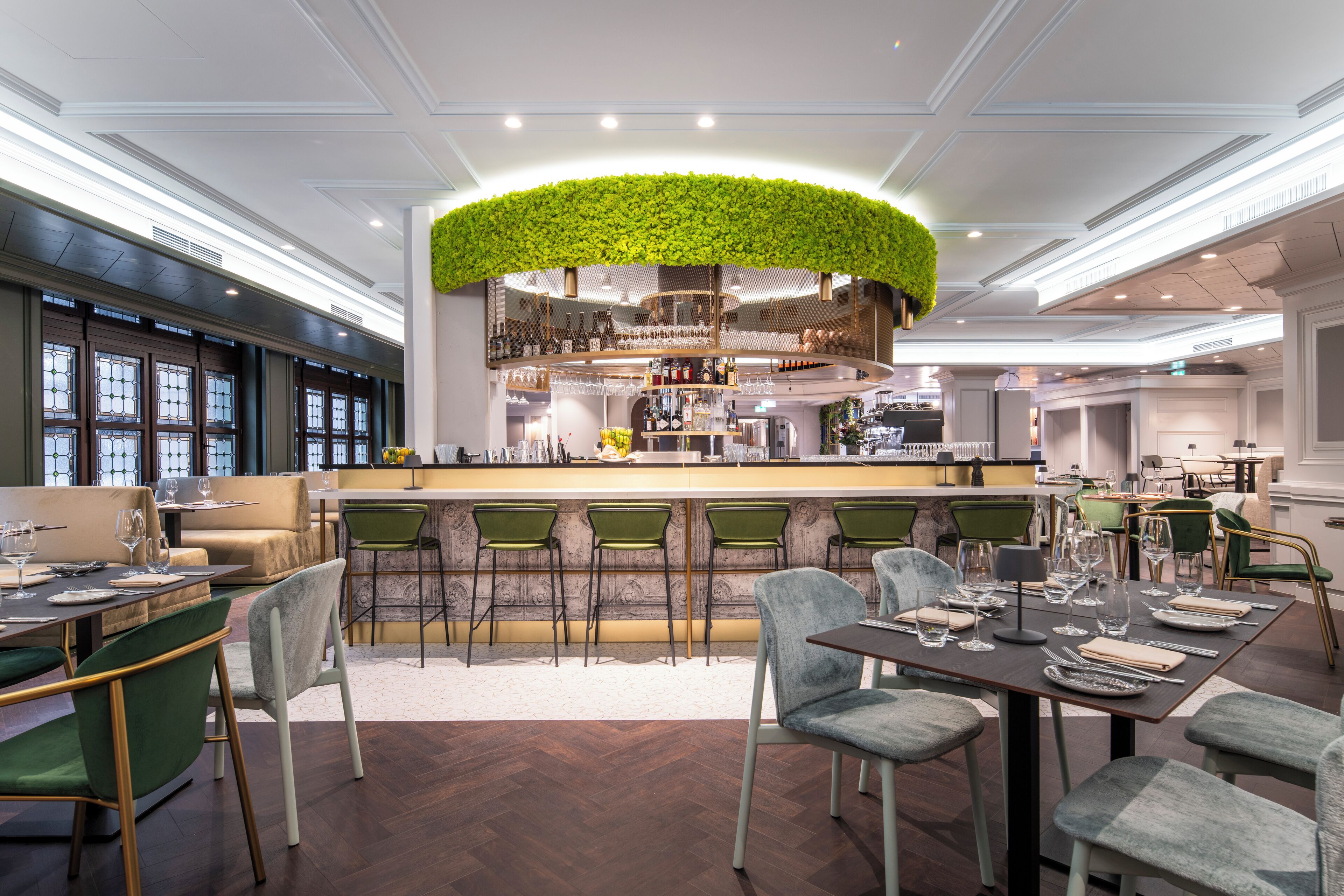 A modern bar area with a green plant ceiling, bar stools, and a variety of bottles and glasses.