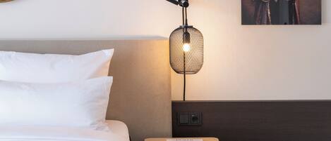 Habitación de hotel con una cama, una mesita de noche de madera con un libro, una lámpara de pared e un retrato enmarcado.