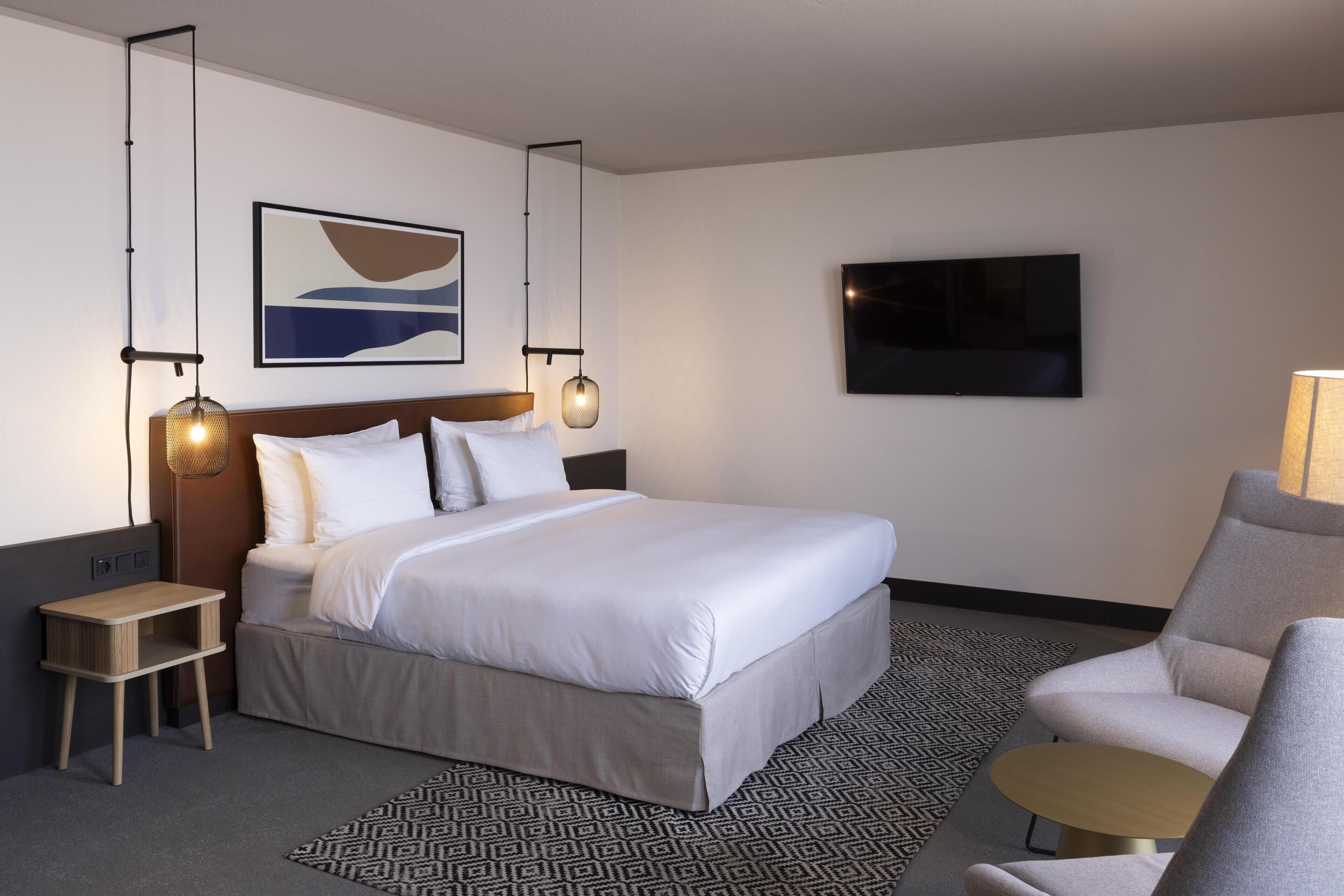 A hotel room with a large bed, a bedside table, a chair, a TV, and a wall-mounted artwork.