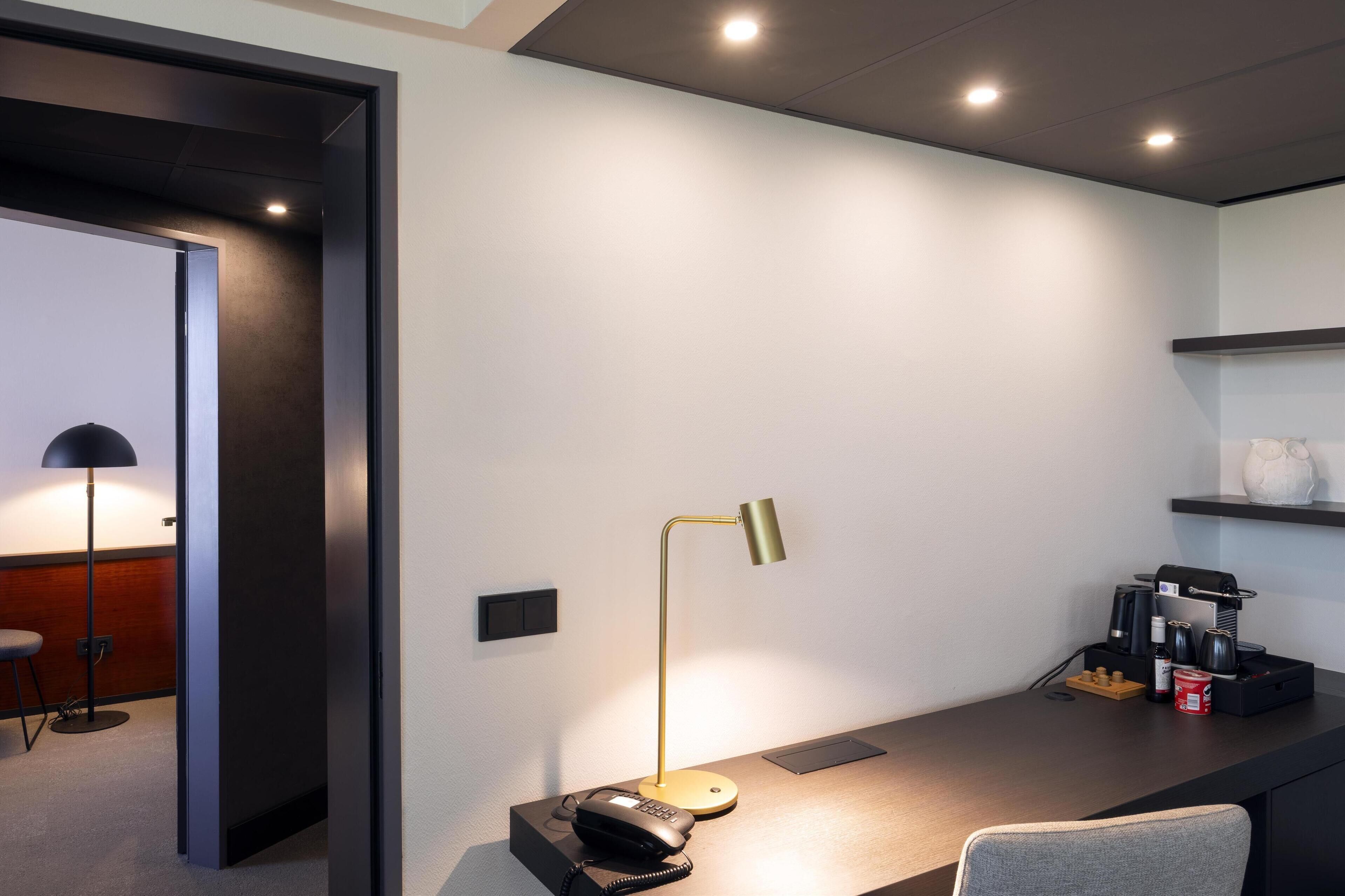 A modern hotel room with a desk, a desk lamp, a telephone, a coffee machine, and a small seating area with a chair and a stool.