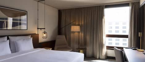 Habitación de hotel con una cama grande, un escritorio, una silla, una lámpara y vistas de edificios a través de la ventana.