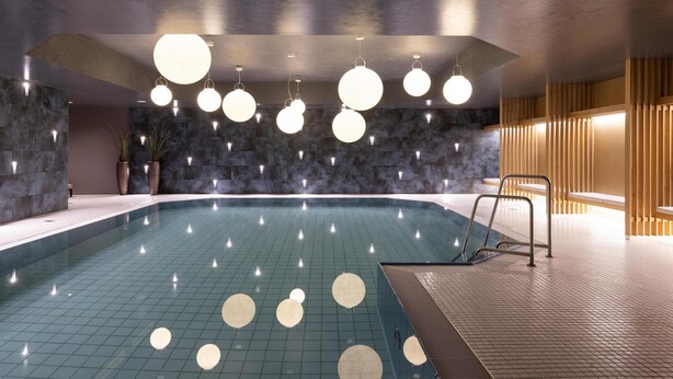 A modern indoor swimming pool with a geometric ceiling design, stone walls, and a wooden seating area. - Scandic Nürnberg Central (Nuremberg)