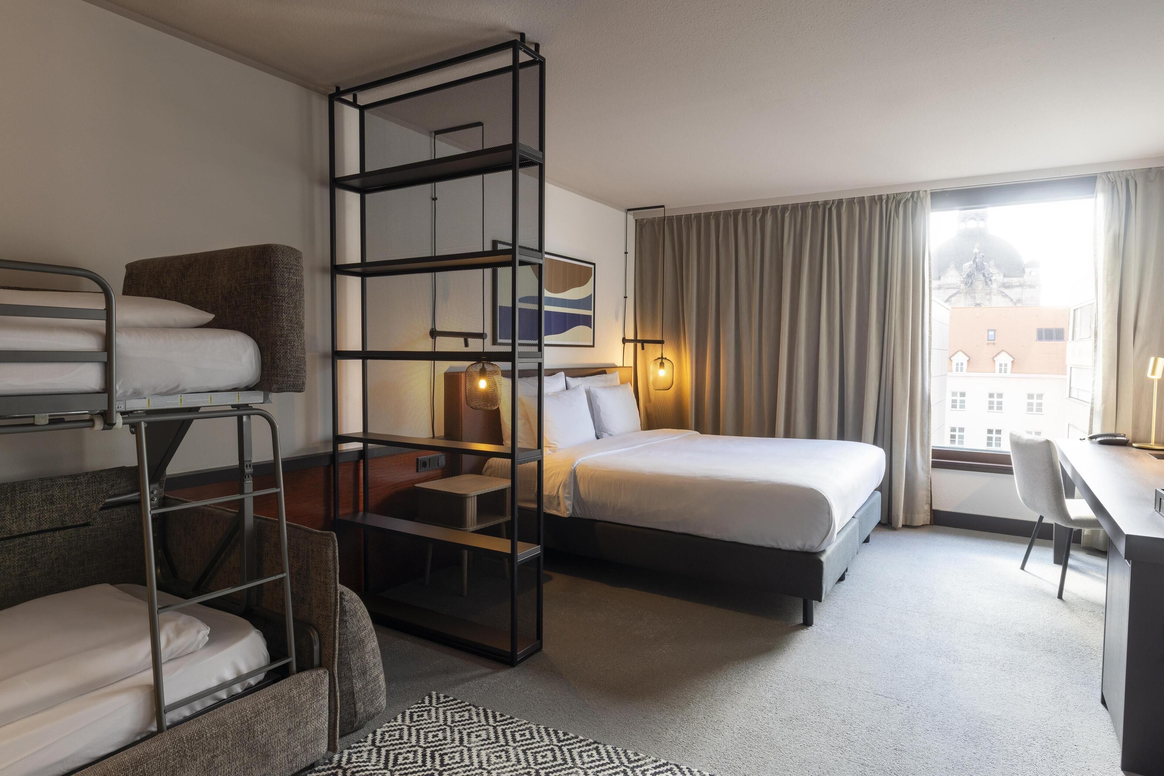 A hotel room with bunk beds, a desk, a bed, and a view of buildings through the window.