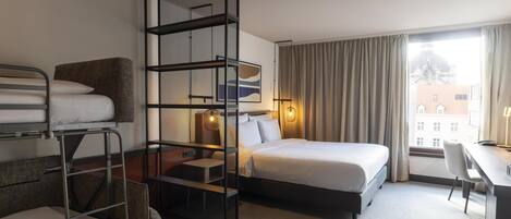 A hotel room with bunk beds, a desk, a bed, and a view of buildings through the window.