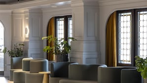 A modern lobby with a seating area featuring a sofa and armchairs, large windows with curtains, and potted plants. - Scandic Nürnberg Central (Nuremberg)