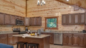 A kitchen with wooden cabinets, granite countertops, a stainless steel dishwasher, and a central island with stools. - Indoor Pool, Theater, Game Room • 10mins to Pigeon Forge | Bluebell by AvantStay (Sevierville)