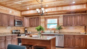 A spacious kitchen with wooden cabinetry, a central island with stools, stainless steel appliances, and a large window providing natural light. - Bluestem & Bluebell by AvantStay | Incredible Views w/ Balcony & Indoor Pool! (Sevierville)