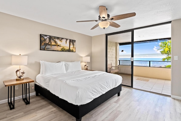 A bedroom with a bed, a ceiling fan, a painting, a nightstand, and a view of the ocean through a glass door.