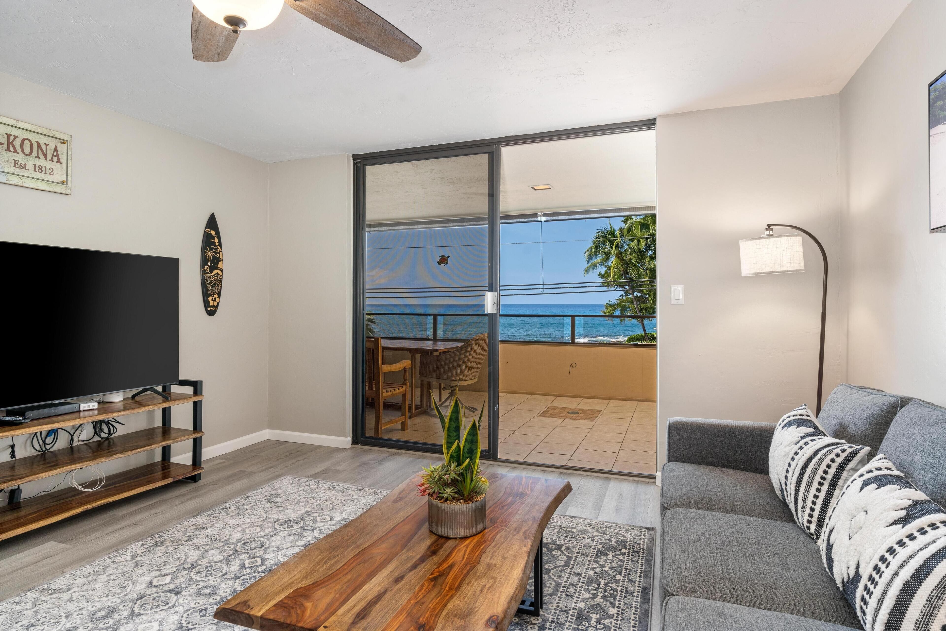 A living room with a flat-screen TV, wooden coffee table, grey sofa, and a balcony with a view of the ocean.