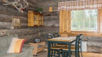Interior rústico de una cabaña con una mesa de comedor de madera, sillas, un sofá, un armario y una ventana con persianas.