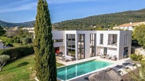 Una casa moderna de dos pisos con piscina, rodeada de vegetación y colinas.