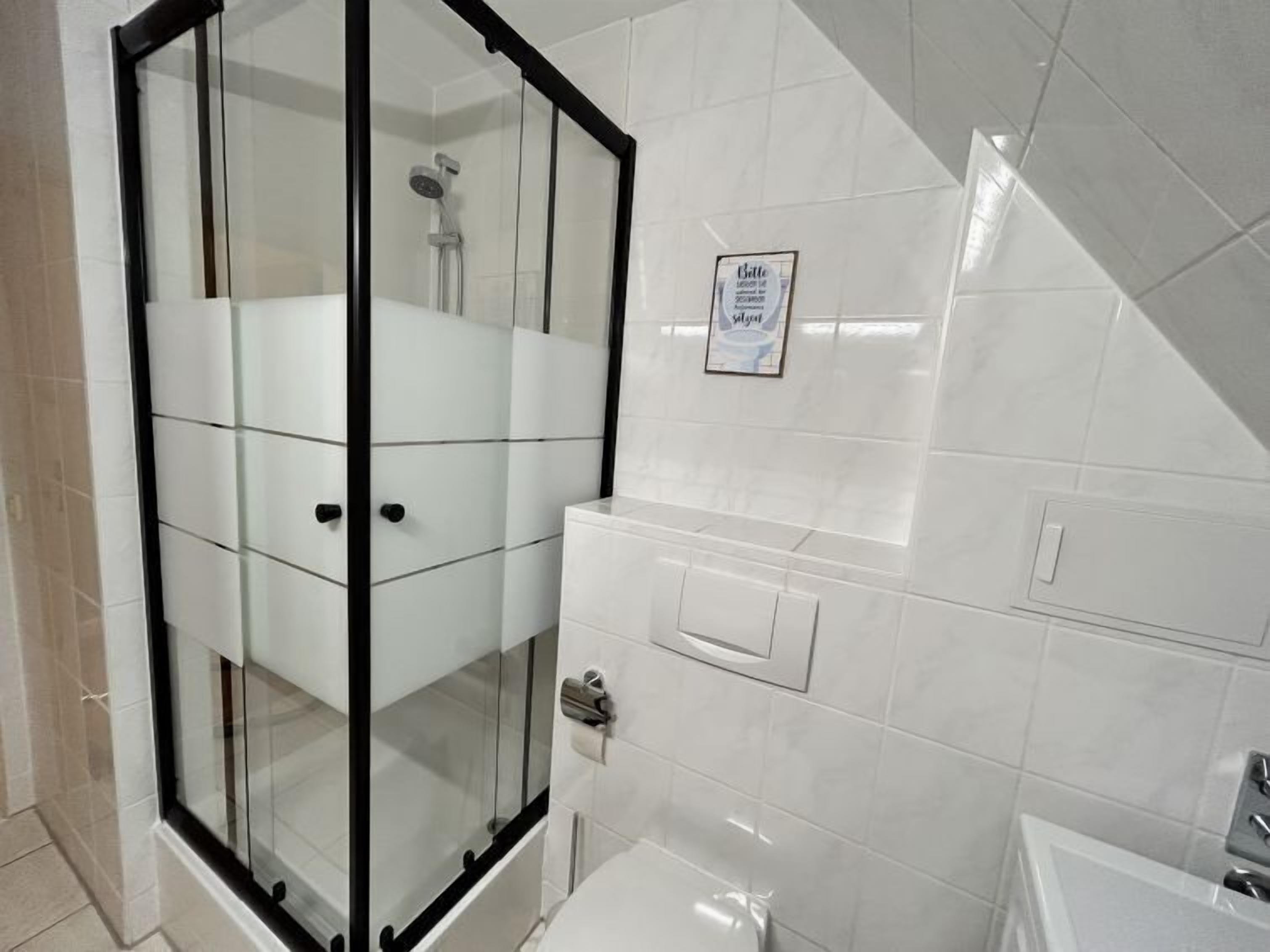 Un baño moderno con ducha acristalada, azulejos blancos y un letrero visible en la pared.