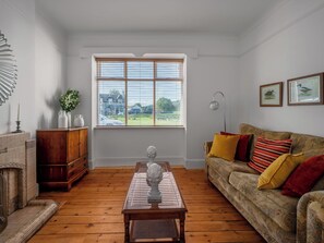 A living room with a wooden floor, a sofa with cushions, a wooden table with a vase, a mirror, and a window with blinds.