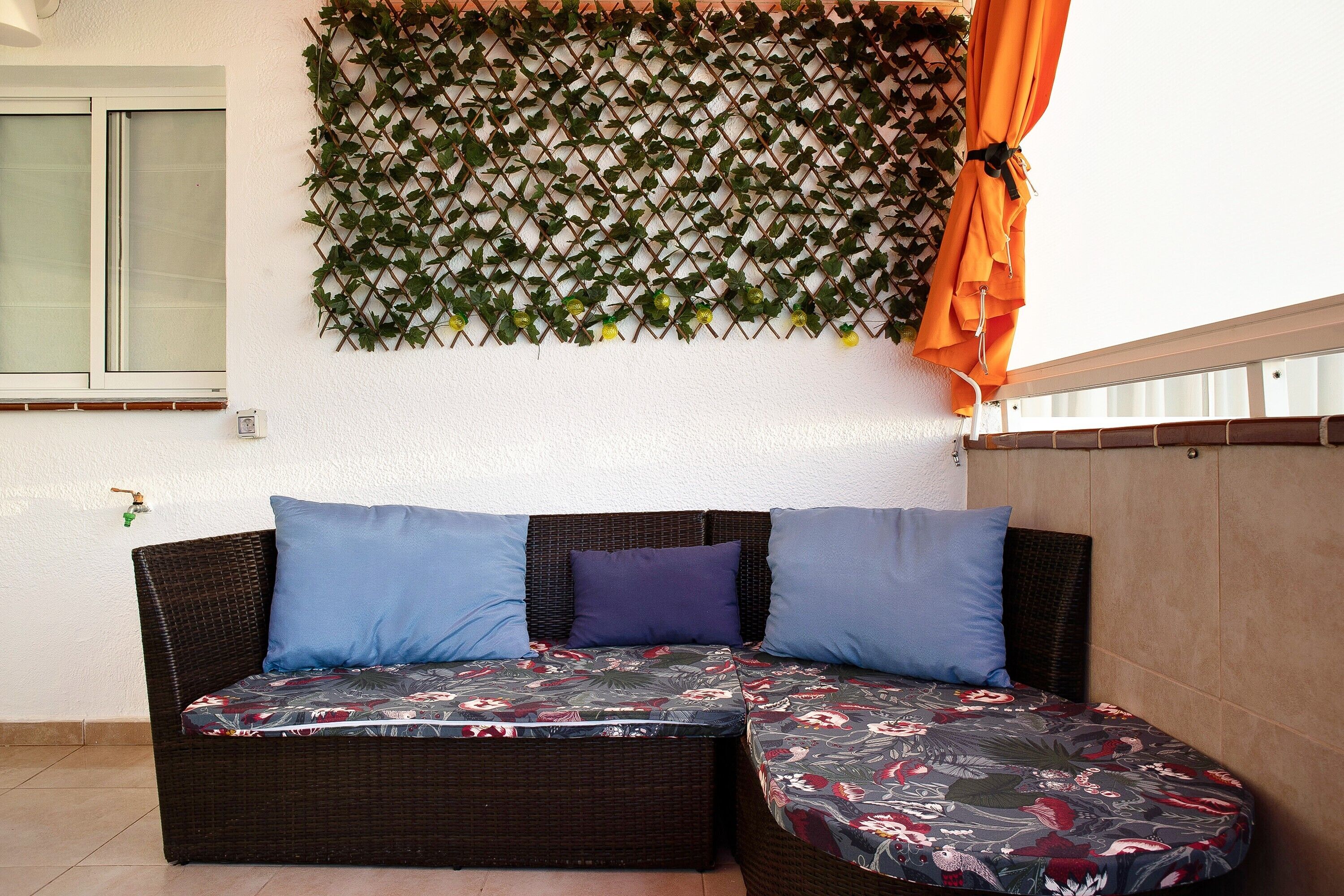 Een rieten bank met bloemenkussens, een muur met een spijlenstructuur begroeid met groen, en een oranje gordijn.