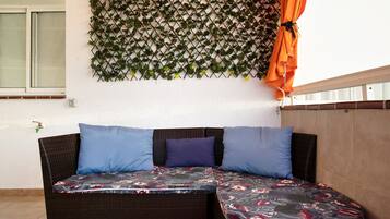 Een rieten bank met bloemenkussens, een muur met een spijlenstructuur begroeid met groen, en een oranje gordijn.