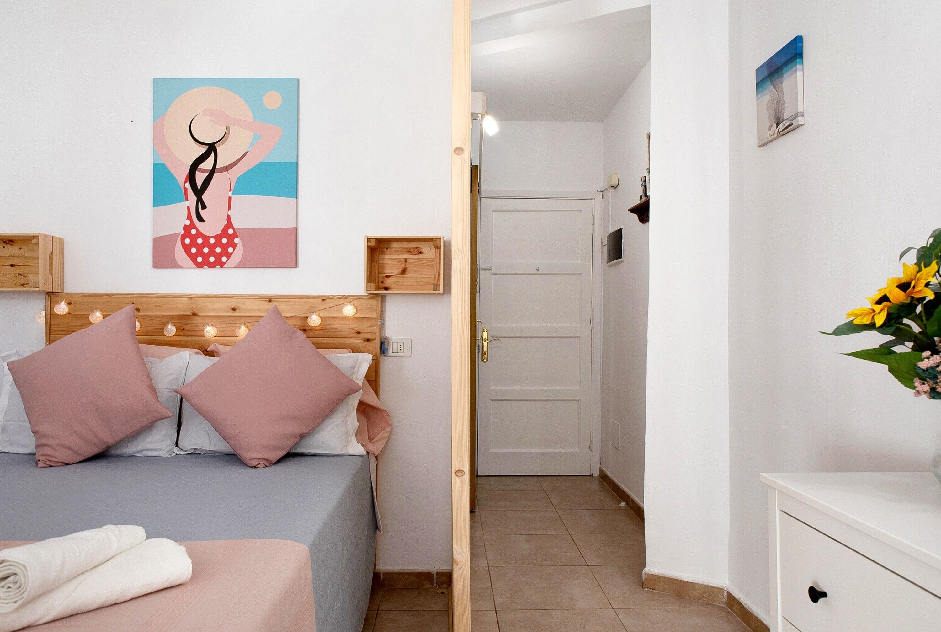 Un dormitorio con cama, almohadas, cabecero, un cuadro, estante de madera, mesita de noche y una imagen colgada en la pared.