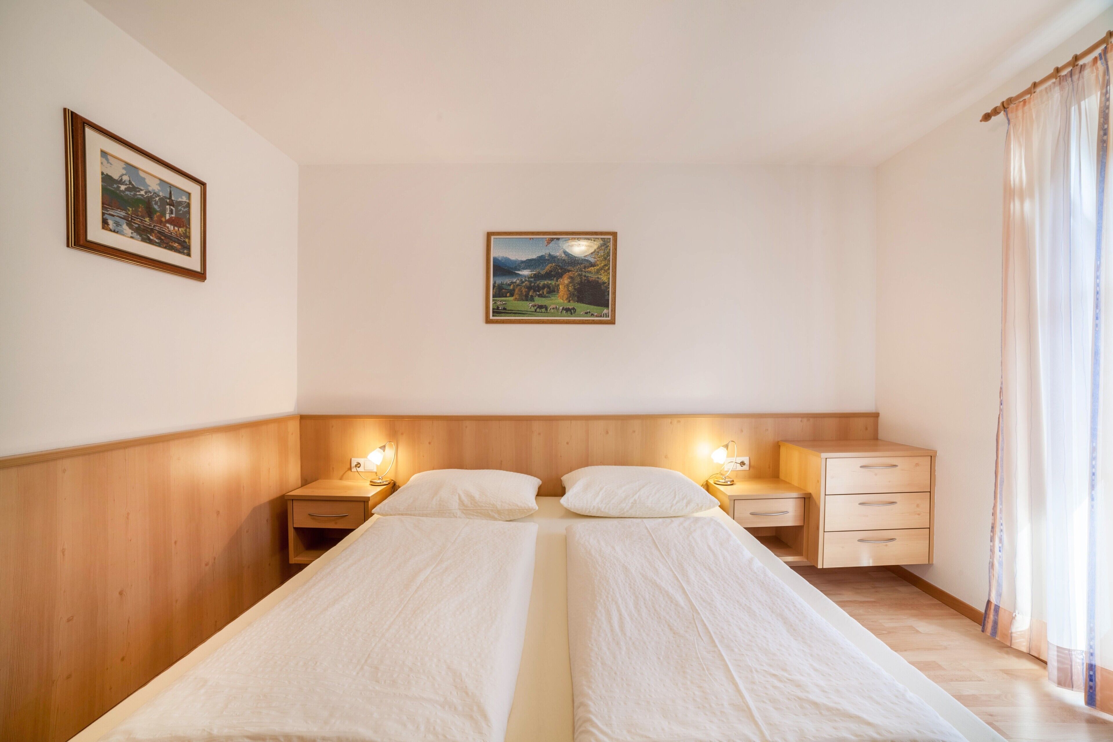 A room with two single beds, wooden headboard, bedside tables with lamps, a small drawer unit, and a framed picture on the wall.