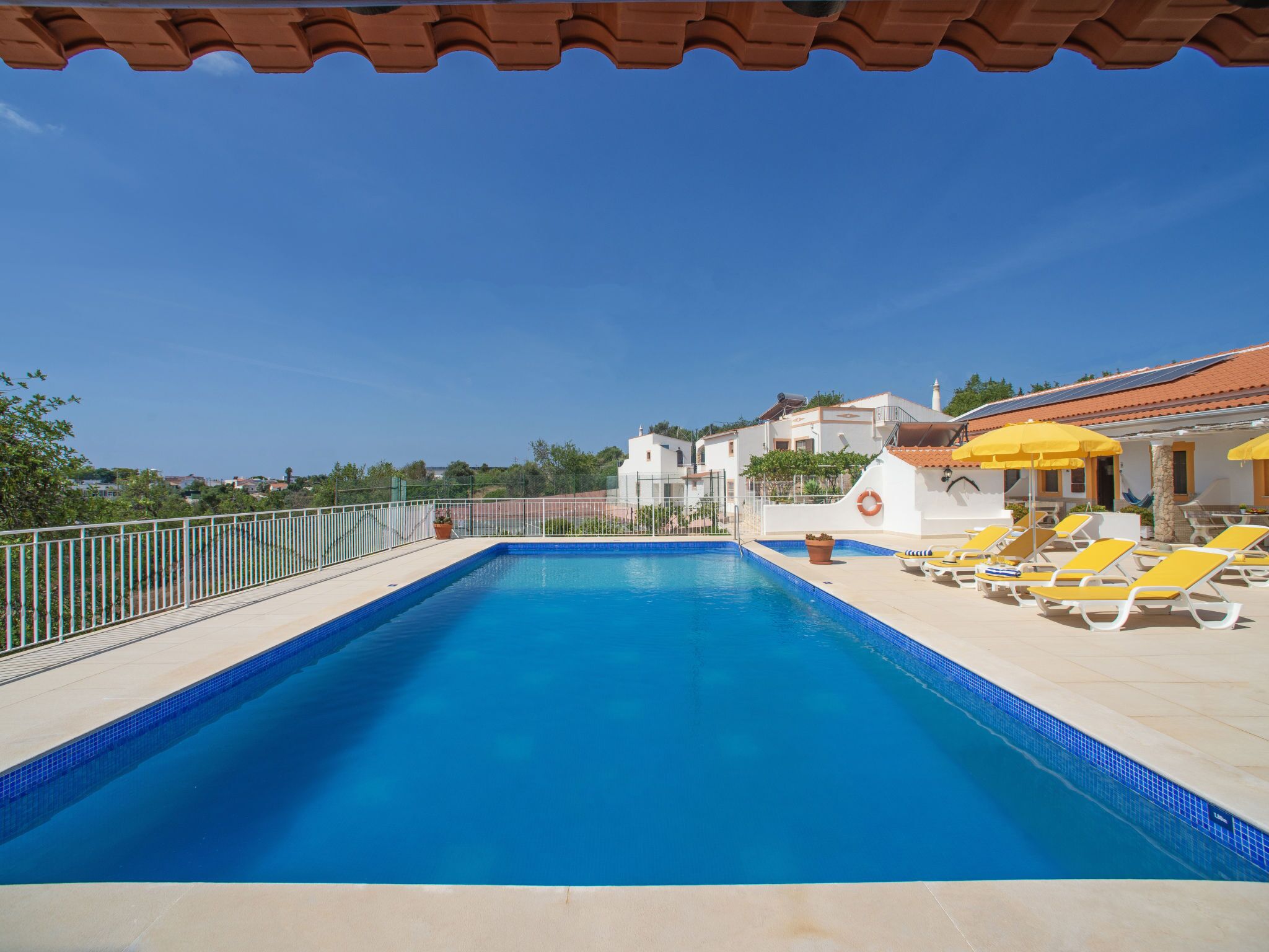 A rectangular swimming pool with a tiled edge, surrounded by a white fence. There are yellow lounge chairs and a yellow umbrella on the right side.