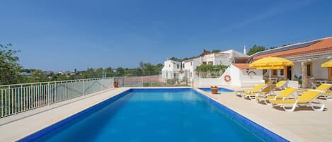 A rectangular swimming pool with a tiled edge, surrounded by a white fence. There are yellow lounge chairs and a yellow umbrella on the right side.
