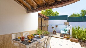 Um pátio externo com uma mesa posta para dois, rodeado por cadeiras brancas, uma parede de pedra e um banco azul.