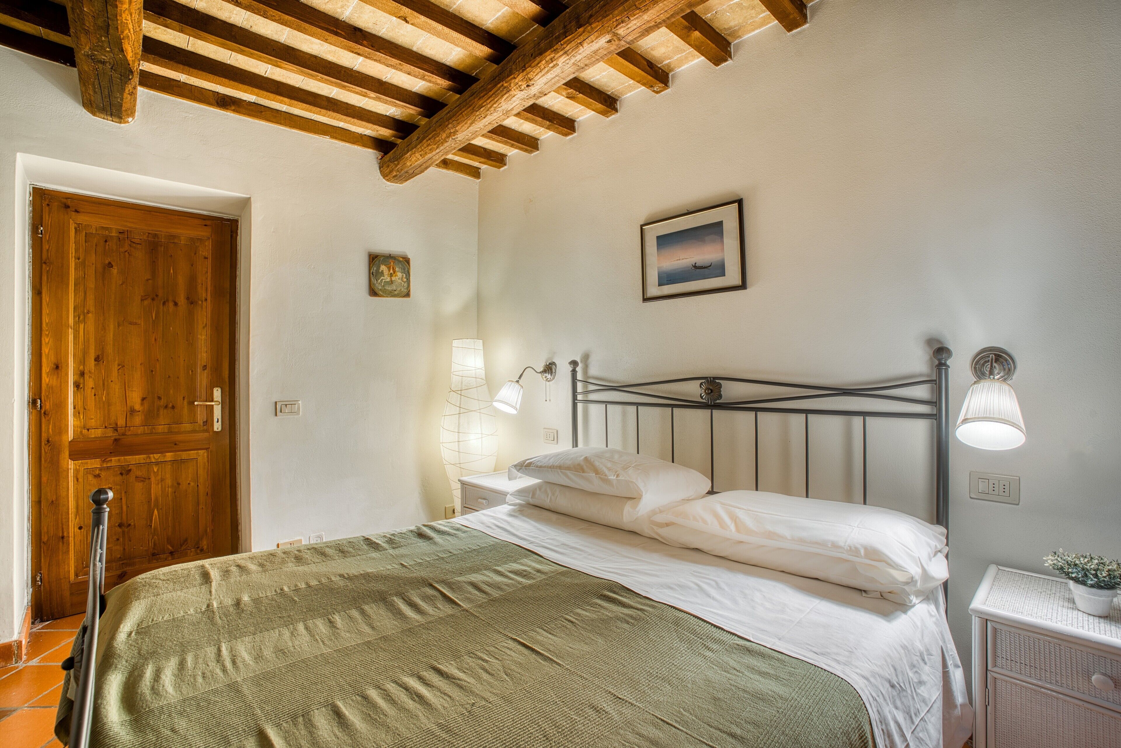 A bedroom with a wooden door, a bed with white linens, a metal headboard, and a small bedside table with a lamp.