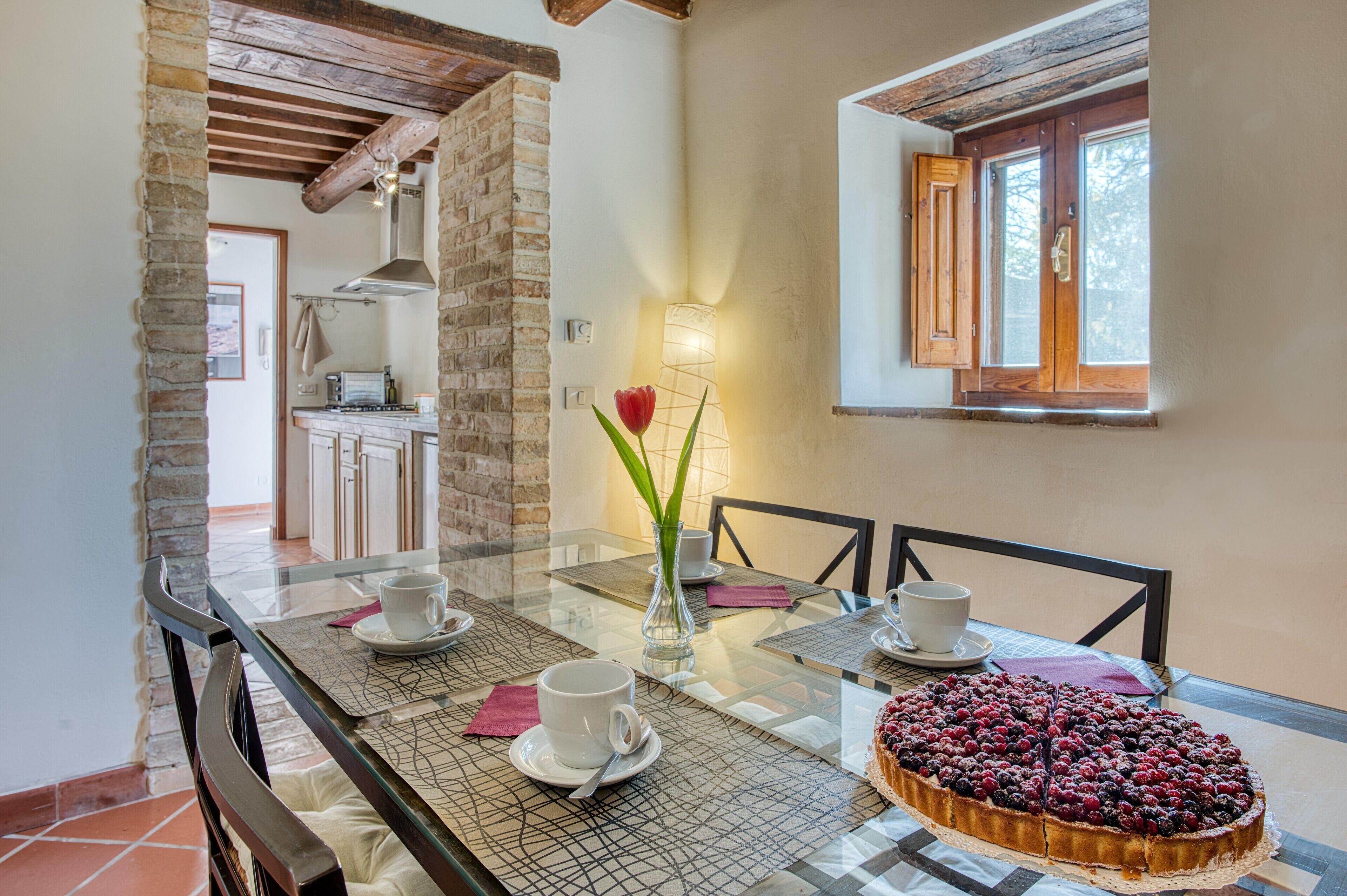 A dining area with a glass table, chairs, a cake, and a vase with a flower.