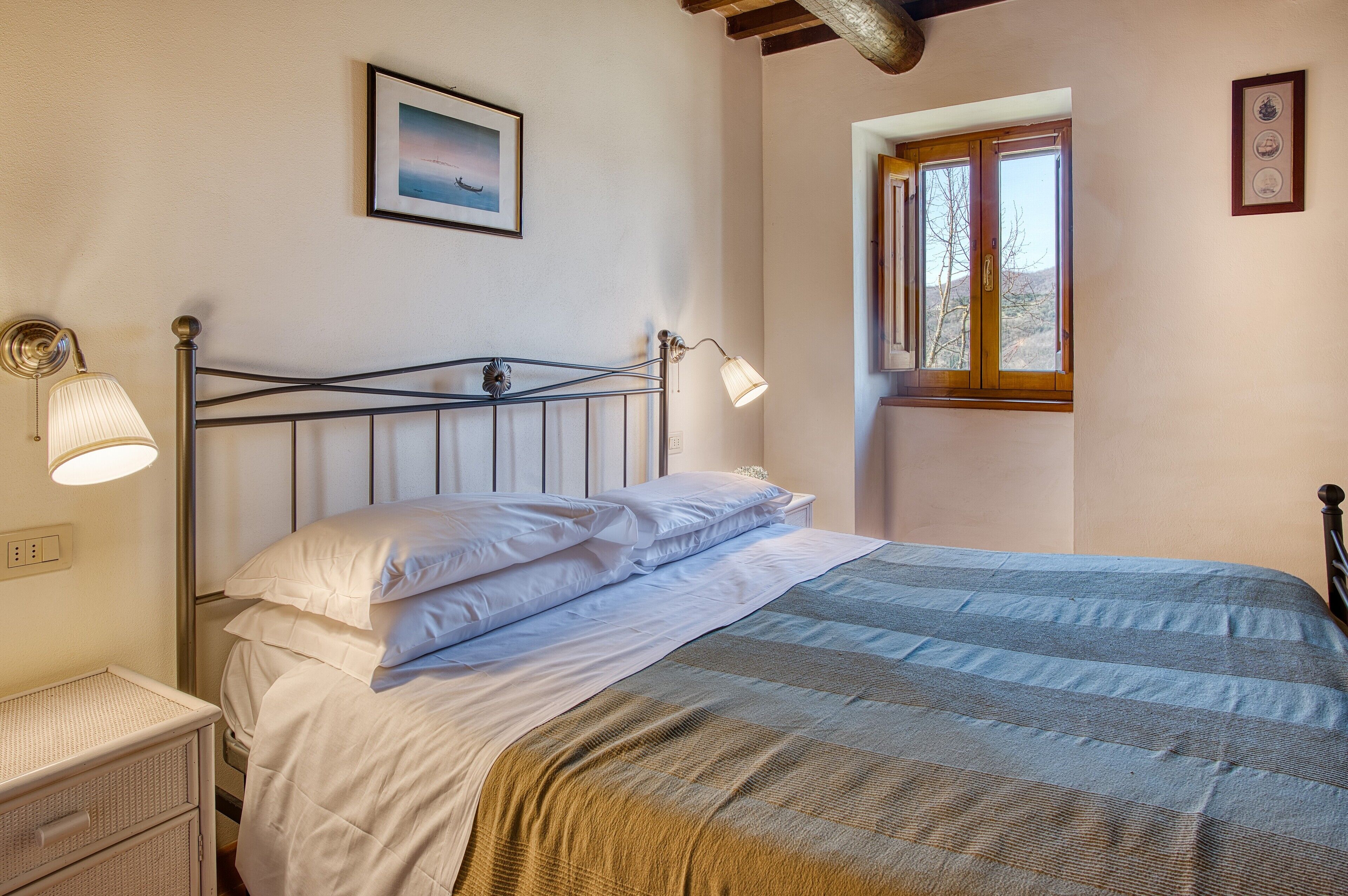 A bed with white linens, a bedside lamp, a framed picture on the wall, and a window with wooden shutters.