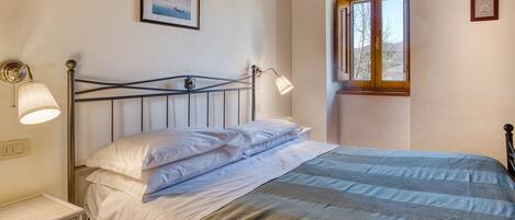 A bed with white linens, a bedside lamp, a framed picture on the wall, and a window with wooden shutters.