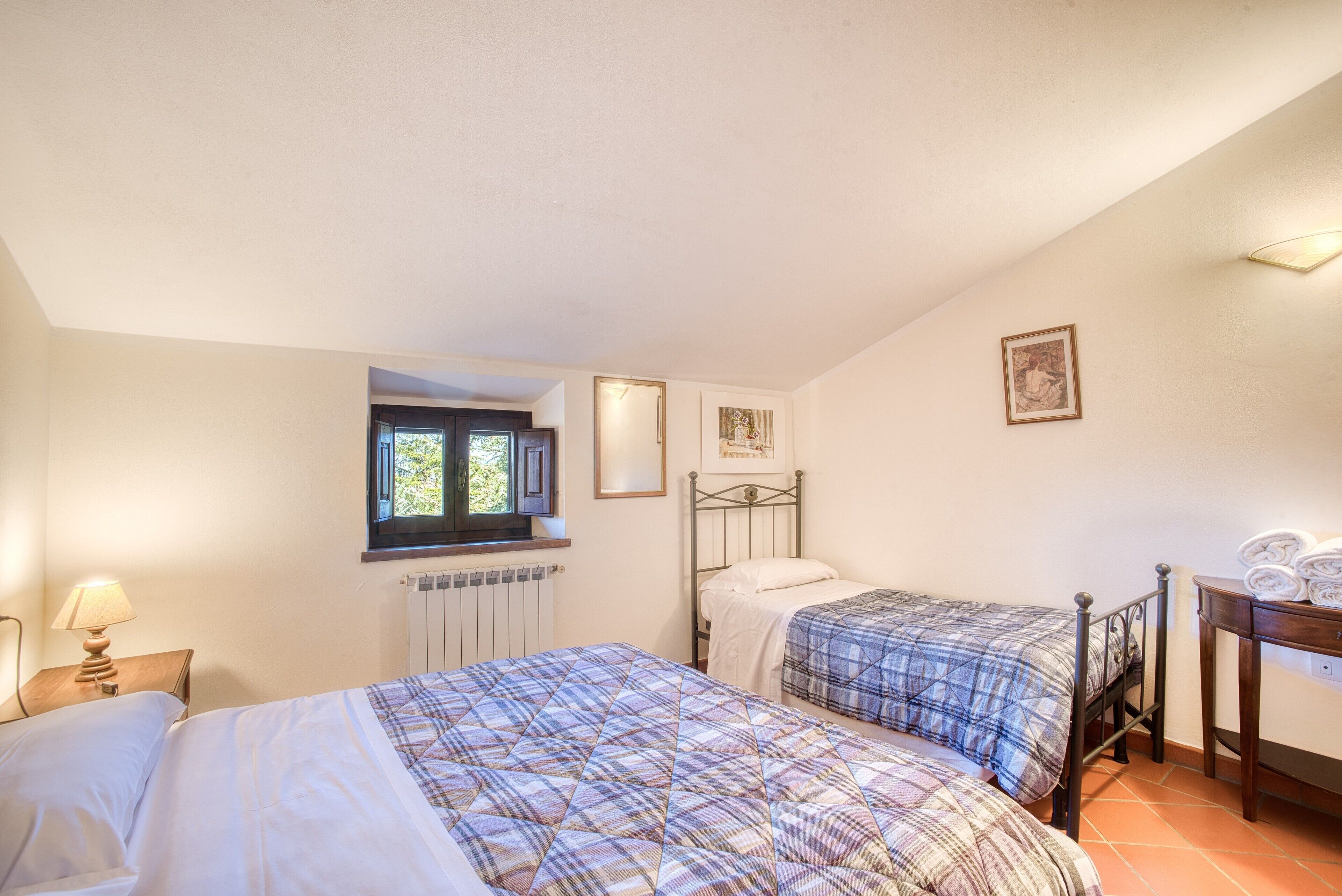 Un dormitorio con dos camas, una ventana, un radiador, una lámpara y un cuadro enmarcado en la pared.