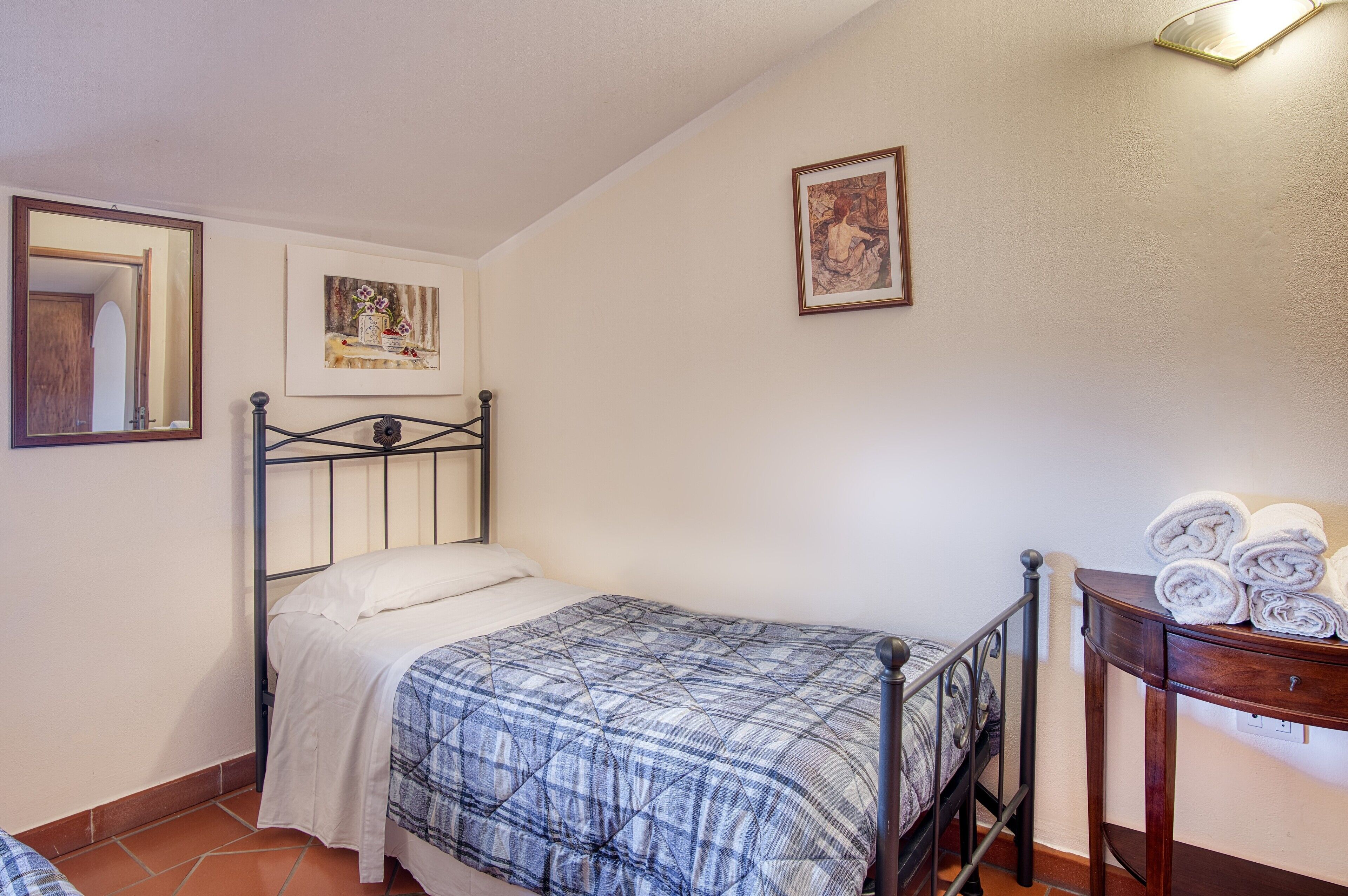 Una cama individual con estructura metálica, una mesita de noche con toallas enrolladas, un espejo sujeto a la pared y dos obras de arte enmarcadas.