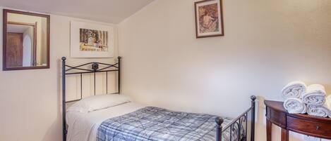 Una cama individual con estructura metálica, una mesita de noche con toallas enrolladas, un espejo sujeto a la pared y dos obras de arte enmarcadas.