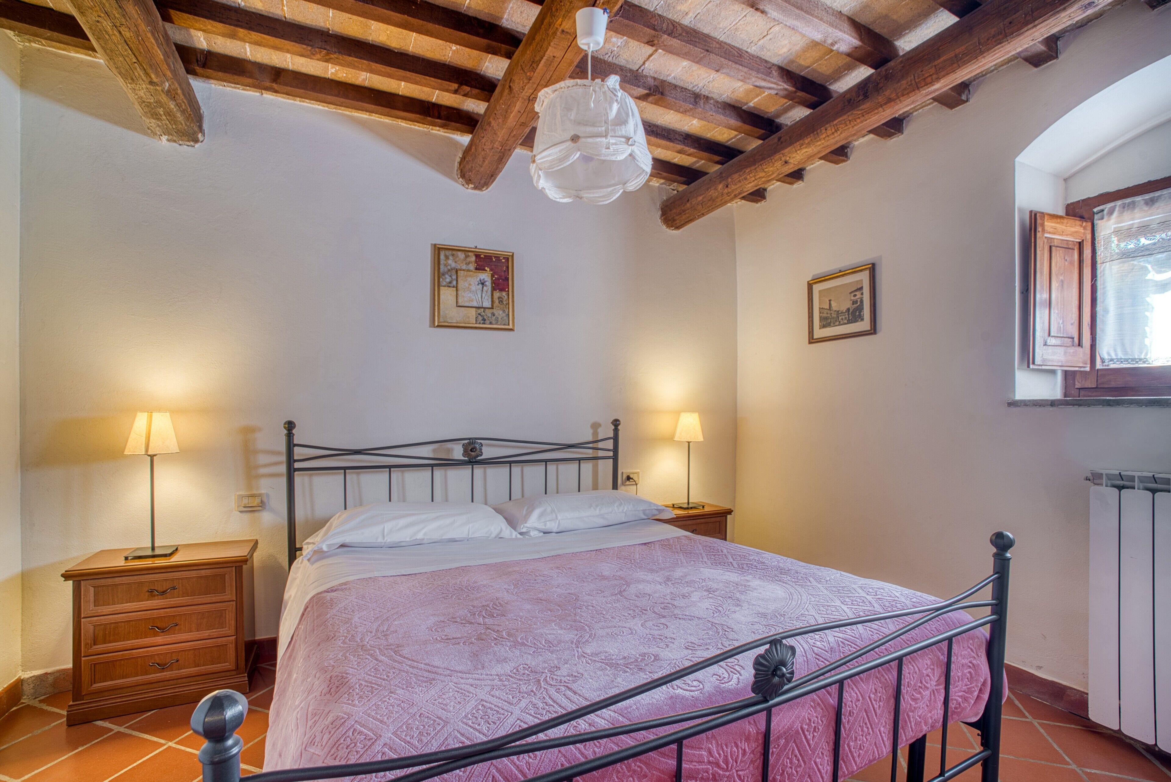 Een slaapkamer met een bed, twee nachtkastjes met lampen, houten balken in het plafond en twee ingekaderde foto's aan de wand.